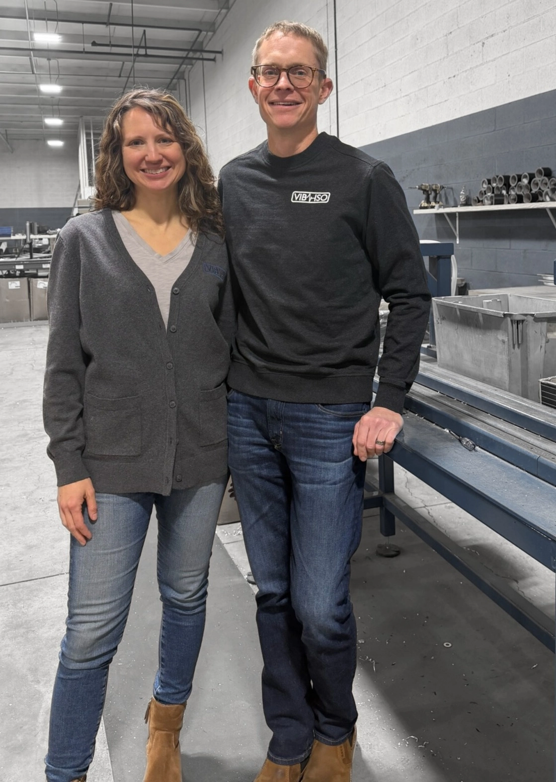 Two people smiling, casually dressed, standing indoors near bleachers.