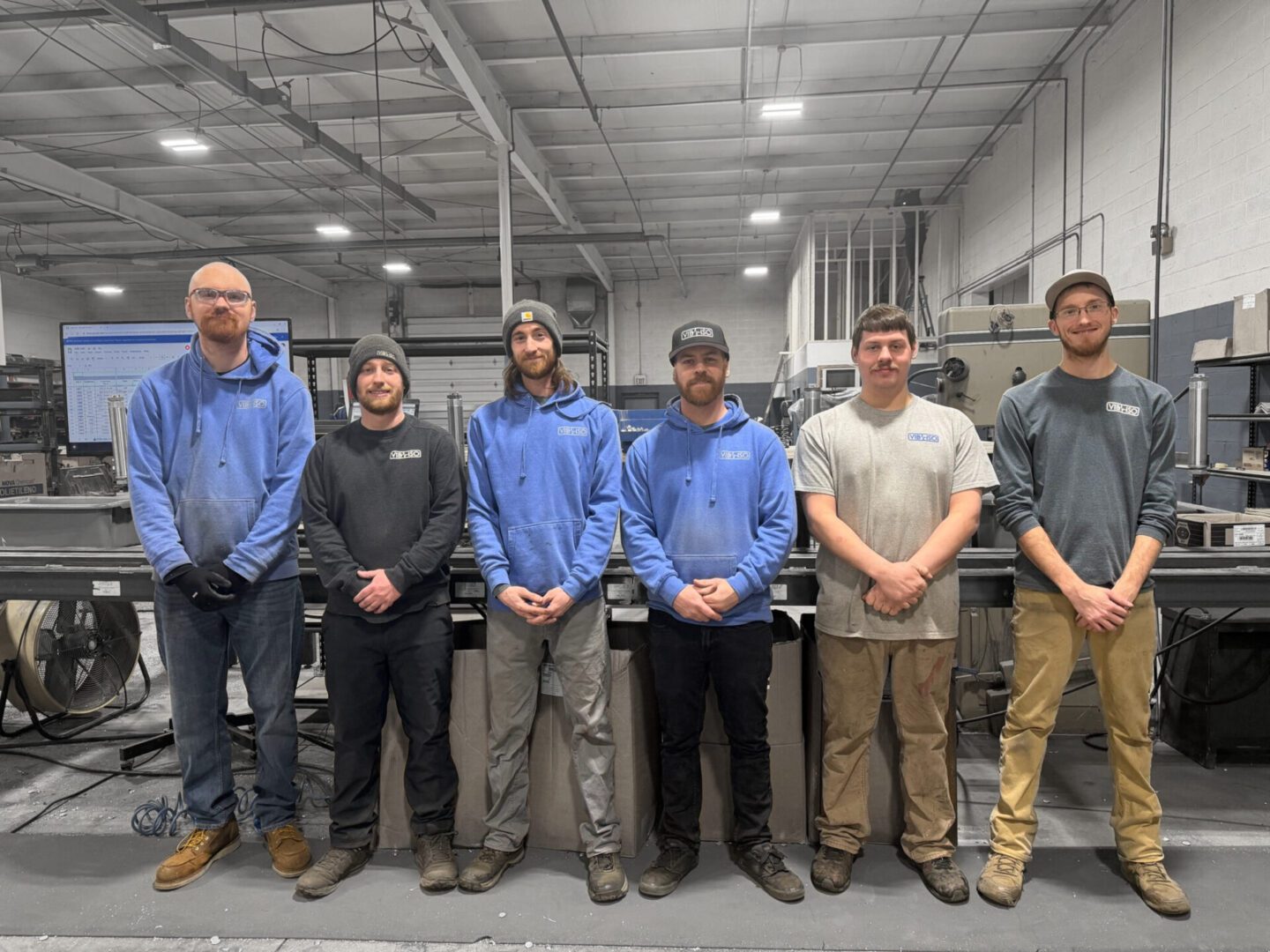 A group of six men standing in a warehouse or industrial setting.